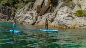 Dos personas practicando paddle surf en Ibiza junto a una costa rocosa de aguas tranquilas